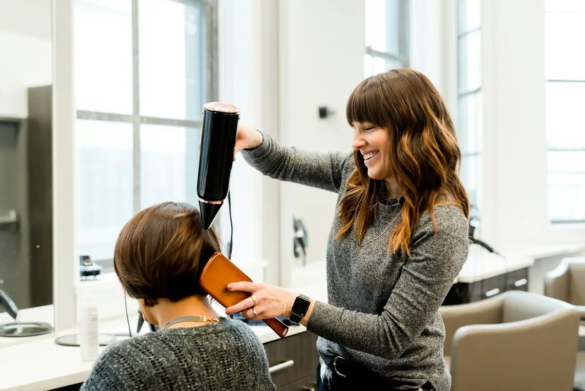 Stylist blow-drying a client's bob and smiling in a bright salon